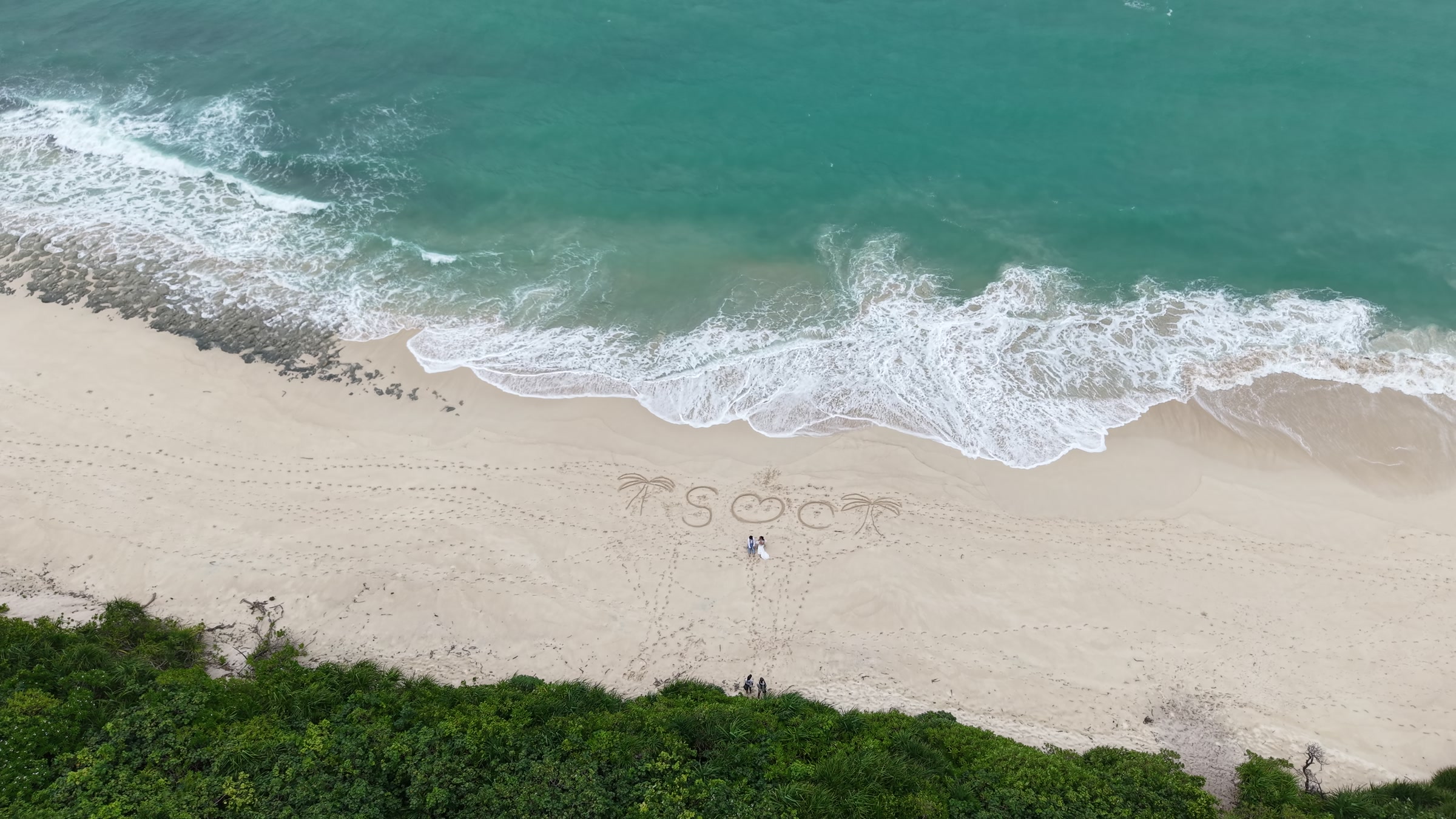 Aerial still from a pre-wedding film in Okinawa: a couple beside a heart drawn in the sand
