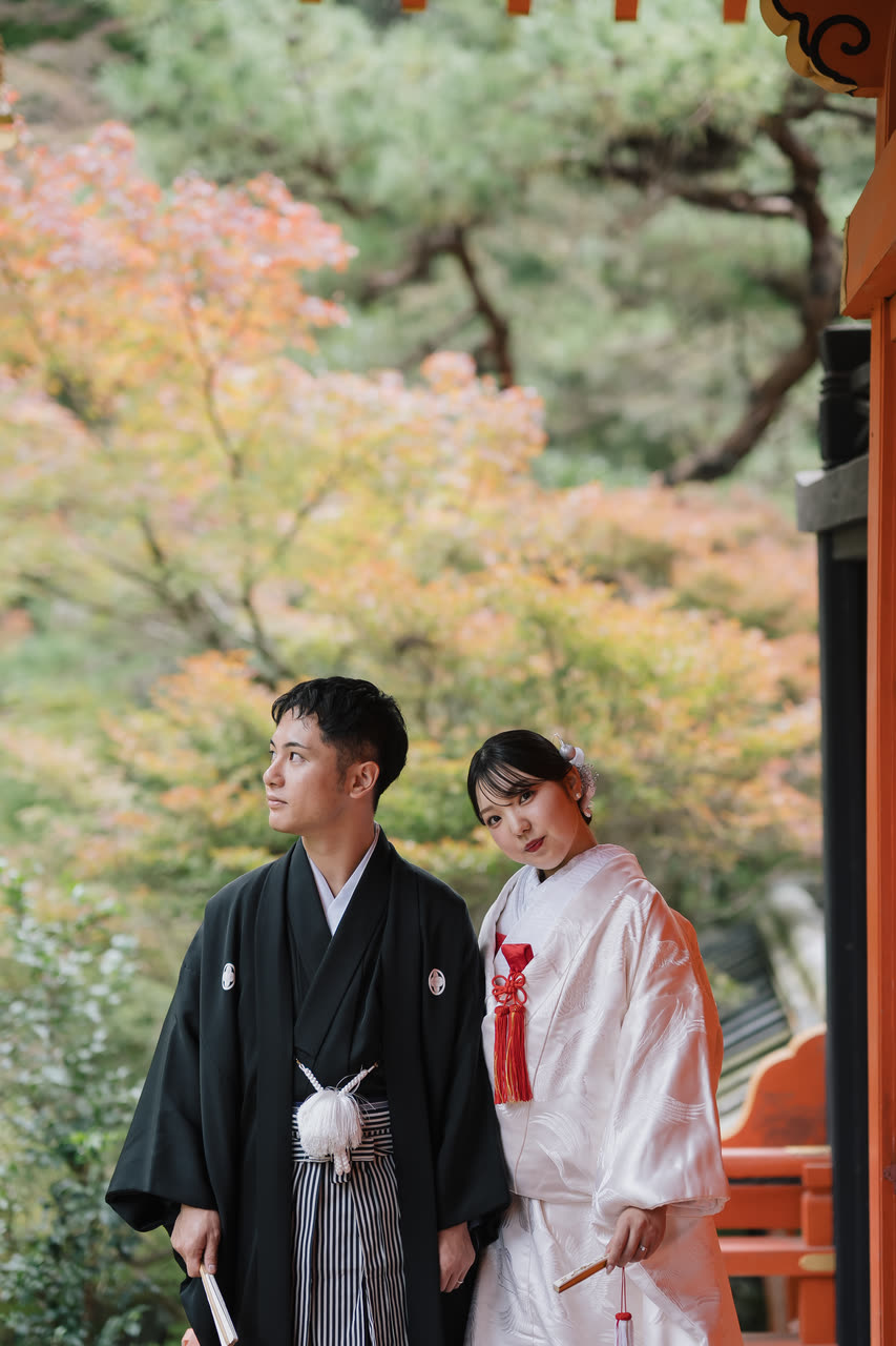 Bride in shiromuku and groom in montsuki standing together among autumn maples in a Kyoto temple garden