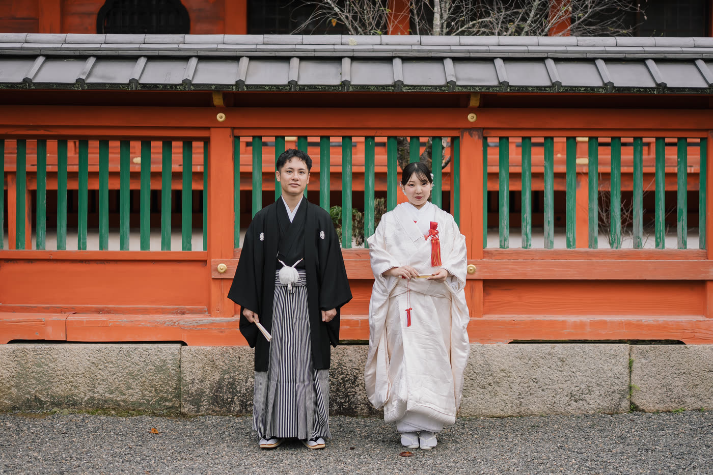 Bride in shiromuku and groom in black montsuki standing together before the vermilion railing of a Kyoto shrine