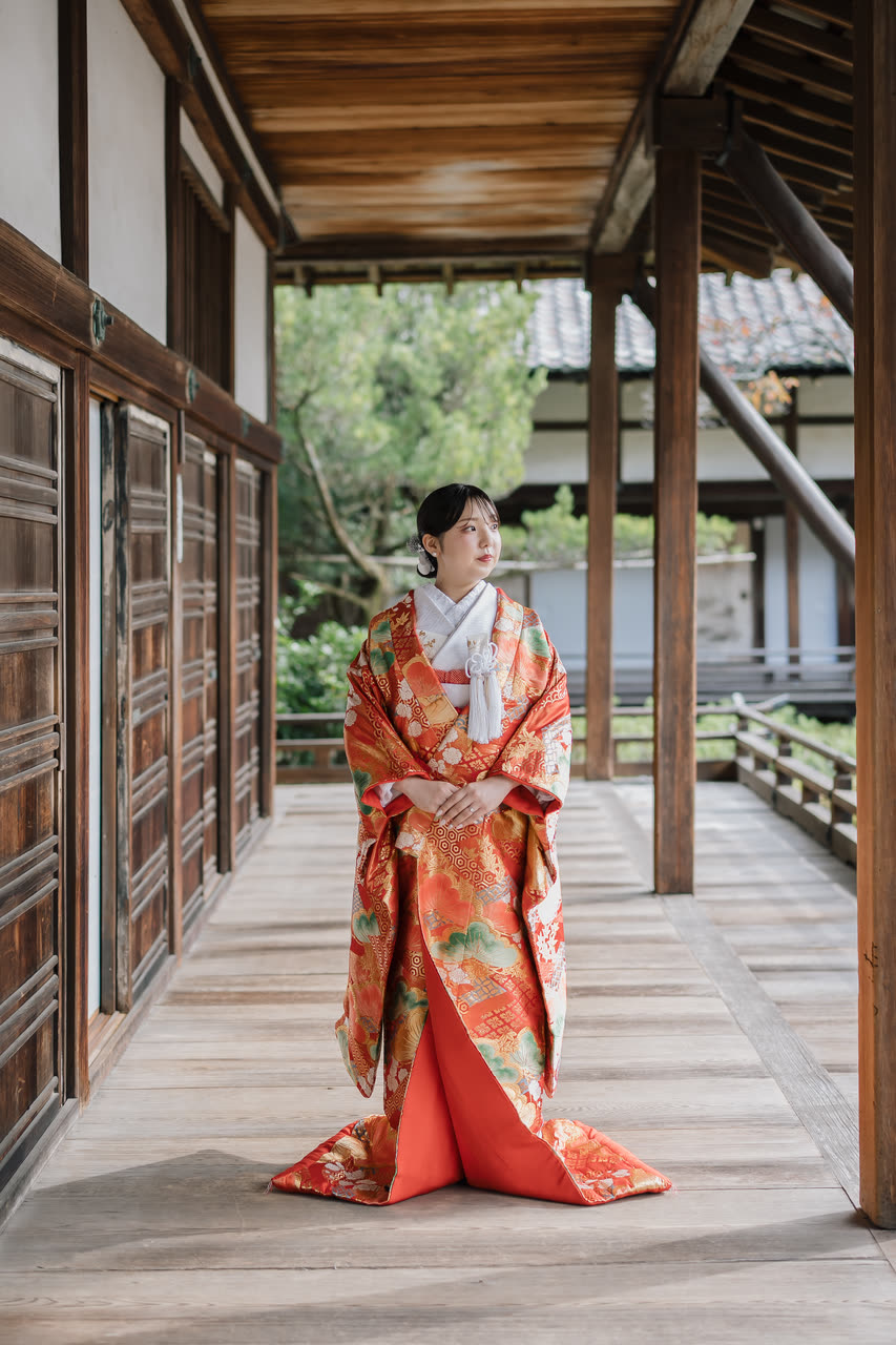 Bride in a red iro-uchikake kimono standing full length in a Kyoto temple corridor