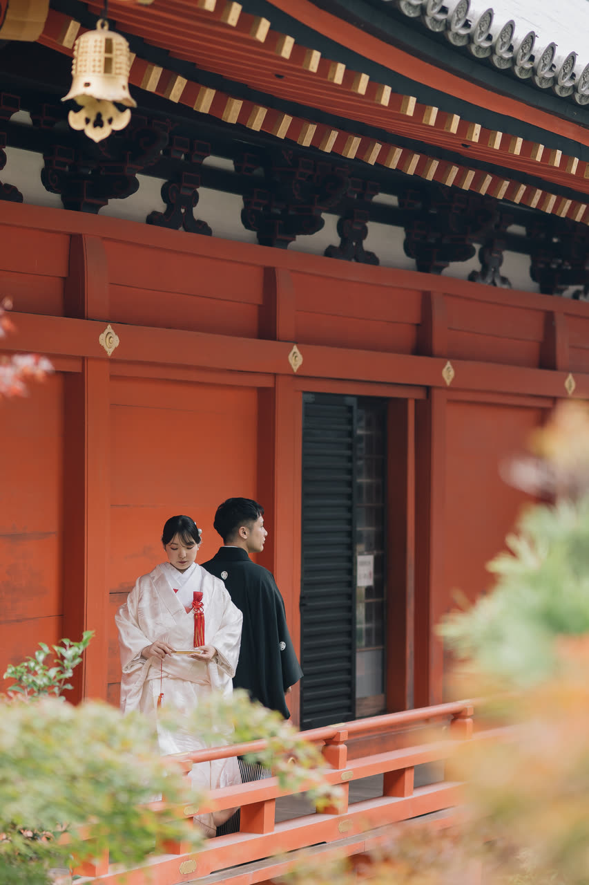 Bride in shiromuku and groom in montsuki on the veranda of a vermilion temple hall framed by autumn maples in Kyoto