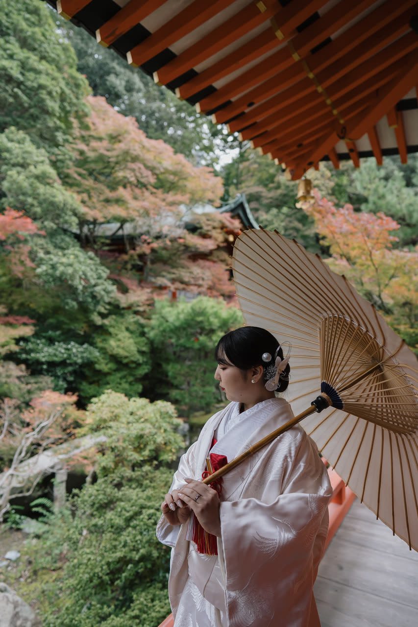 Bride in shiromuku holding a wagasa parasol in profile with autumn maples behind her in Kyoto