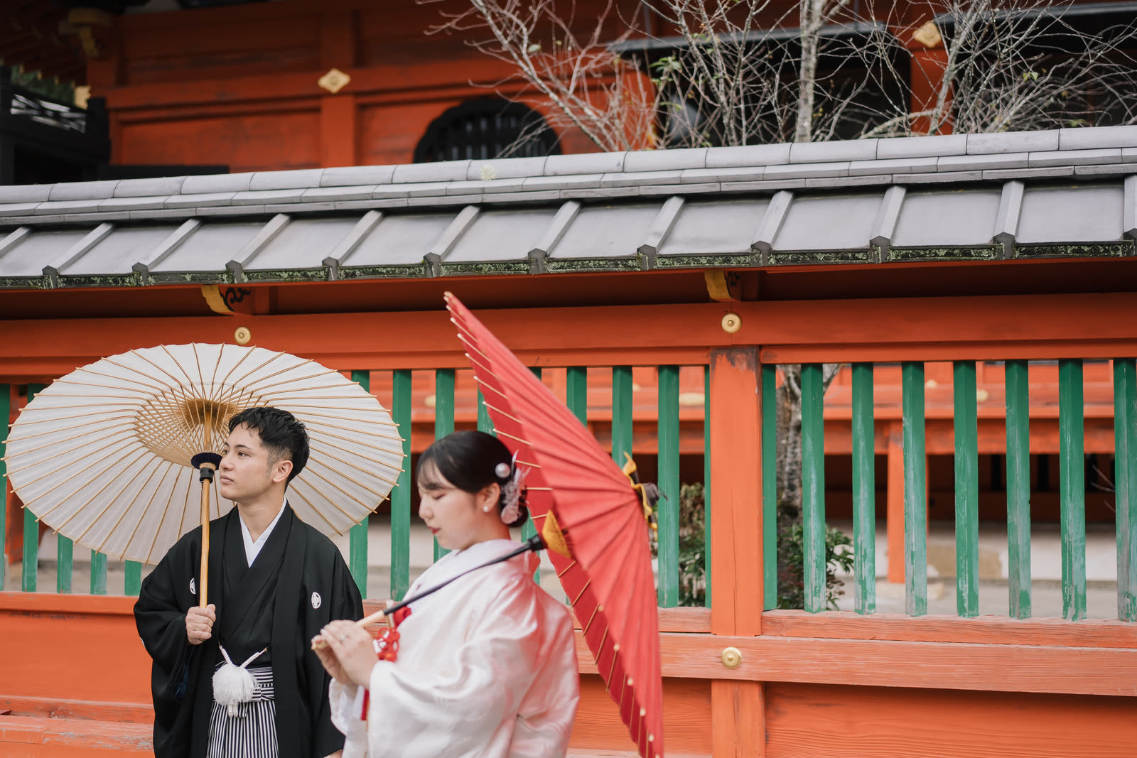Bride and groom holding wagasa parasols beside the vermilion railing of a Kyoto shrine