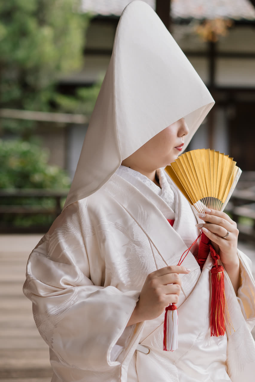 Bride in a white shiromuku kimono and wataboshi hood holding a gold folding fan