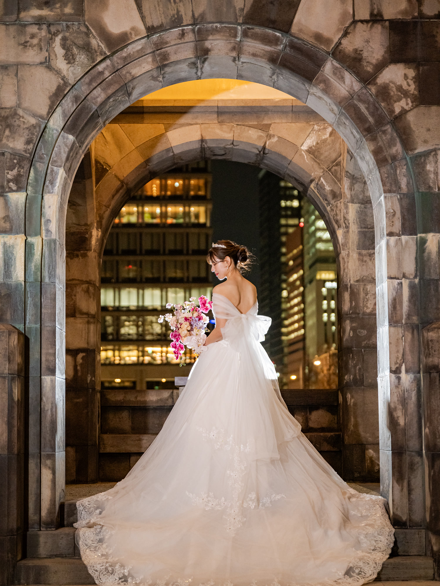 Bride in a long gown beneath a stone arch with Tokyo lights behind her at night