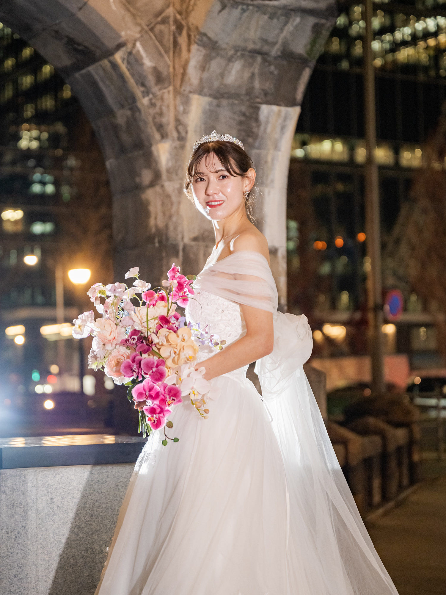 Bride with a bouquet under a stone archway, Tokyo city lights in the background